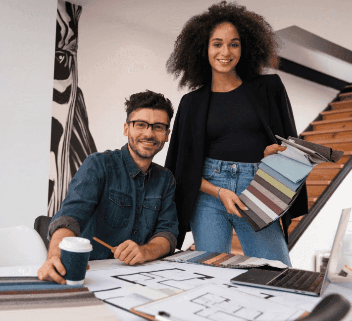 Two interior designers smiling at a desk with floor plans and fabric samples, one standing and holding a swatch book while the other sits with a coffee cup and pencil.