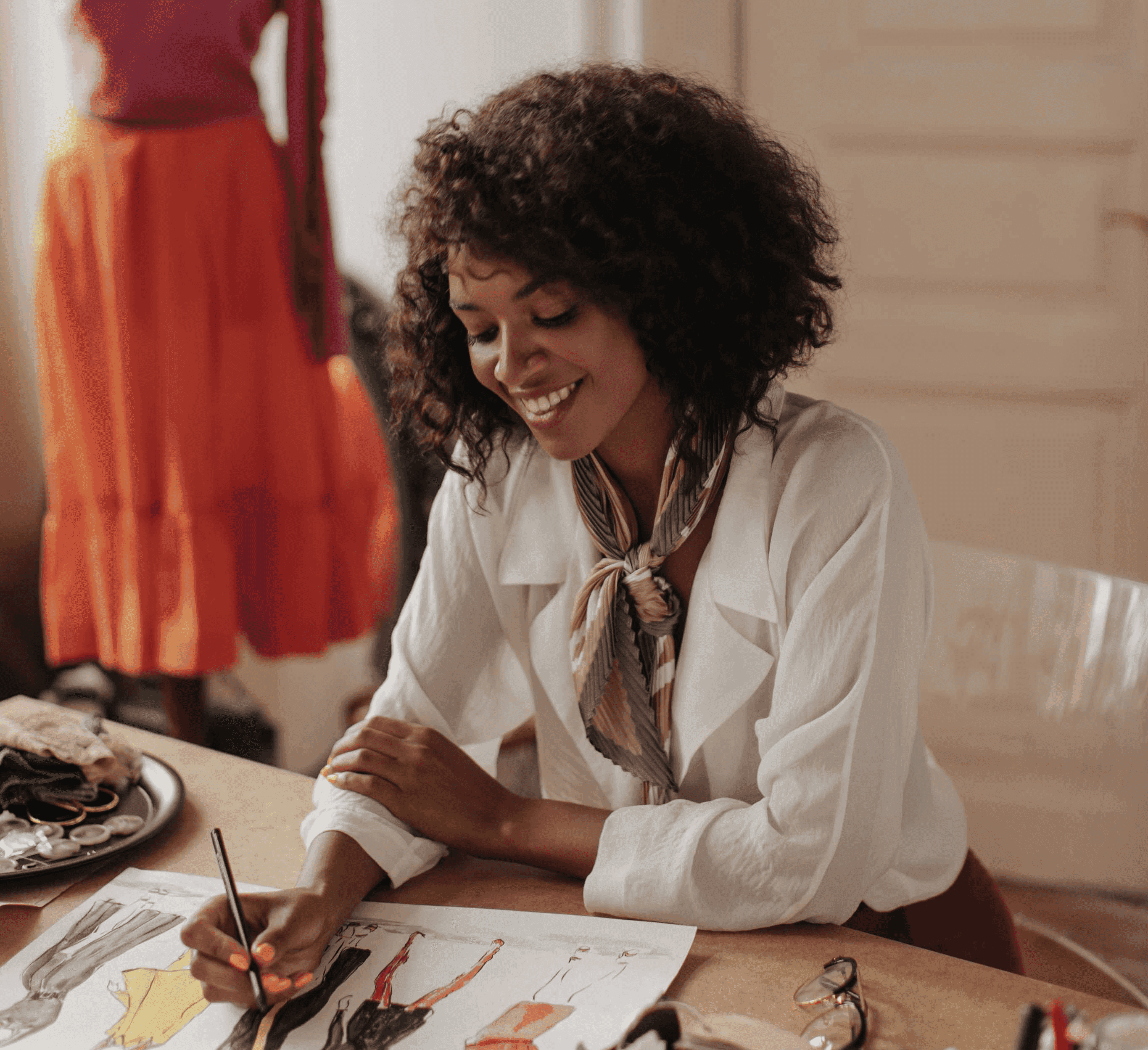 Fashion designer smiling while sketching clothing designs at a desk, with a mannequin wearing a pink top and orange skirt in the background.